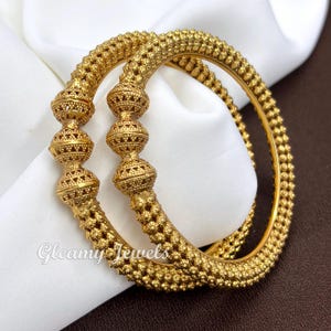 May include: Two gold bangles featuring a textured beaded surface and three prominent, spherical, filigree-style centre beads. The jewellery displays a metallic yellow finish with intricate detailing. The text Gleamy Jewels is visible.