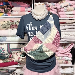 May include: A mannequin displays a patchwork crossbody bag with a variety of fabric patterns in pink, blue, green, and cream. The mannequin wears a dark blue t-shirt with white text and a pink tulle skirt, accessorized with a pearl necklace.