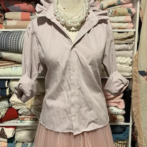 May include: A light pink button-down shirt with a small checkered pattern, a ruffled collar, and rolled-up sleeves. The shirt is paired with a pink tulle skirt. A multi-strand pearl necklace is around the neck. The text "Small Calvin Klein" is at the bottom.