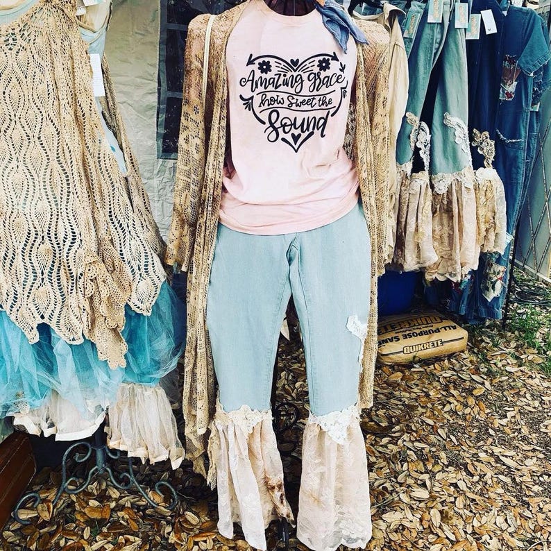 Puede incluir: Una camiseta rosa con un gr&aacute;fico de coraz&oacute;n negro que dice "Amazing Grace How Sweet the Sound". La camiseta se combina con jeans azul claro con ribete de encaje y una chaqueta de punto beige.
