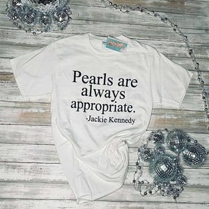 May include: White t-shirt with black text that reads "Pearls are always appropriate. -Jackie Kennedy"