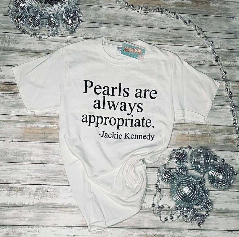 May include: White t-shirt with black text that reads "Pearls are always appropriate. -Jackie Kennedy"