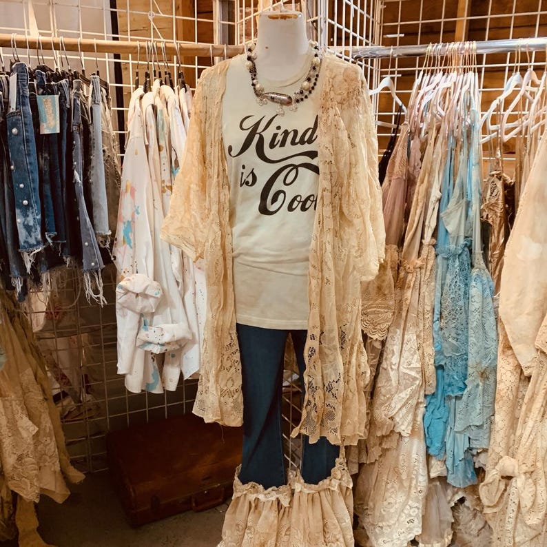 Puede incluir: Una camiseta blanca con el texto "Kind is Cool" impreso en ella. La camiseta la lleva puesta un maniqu&iacute; con una chaqueta de encaje y vaqueros. El maniqu&iacute; est&aacute; de pie delante de una percha de ropa.