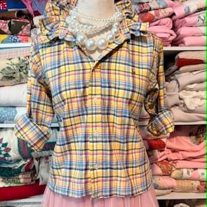 K&ouml;nnte beinhalten: Ein gelb, blau und rosa kariertes Button-Down-Hemd mit hochgekrempelten &Auml;rmeln wird auf einer Schaufensterpuppe pr&auml;sentiert. Das Hemd hat einen R&uuml;schenkragen und ein kleines Logo. Eine mehrreihige Perlenkette wird getragen. Das Wort "Small" ist auf dem rosa Rock aufgedruckt.