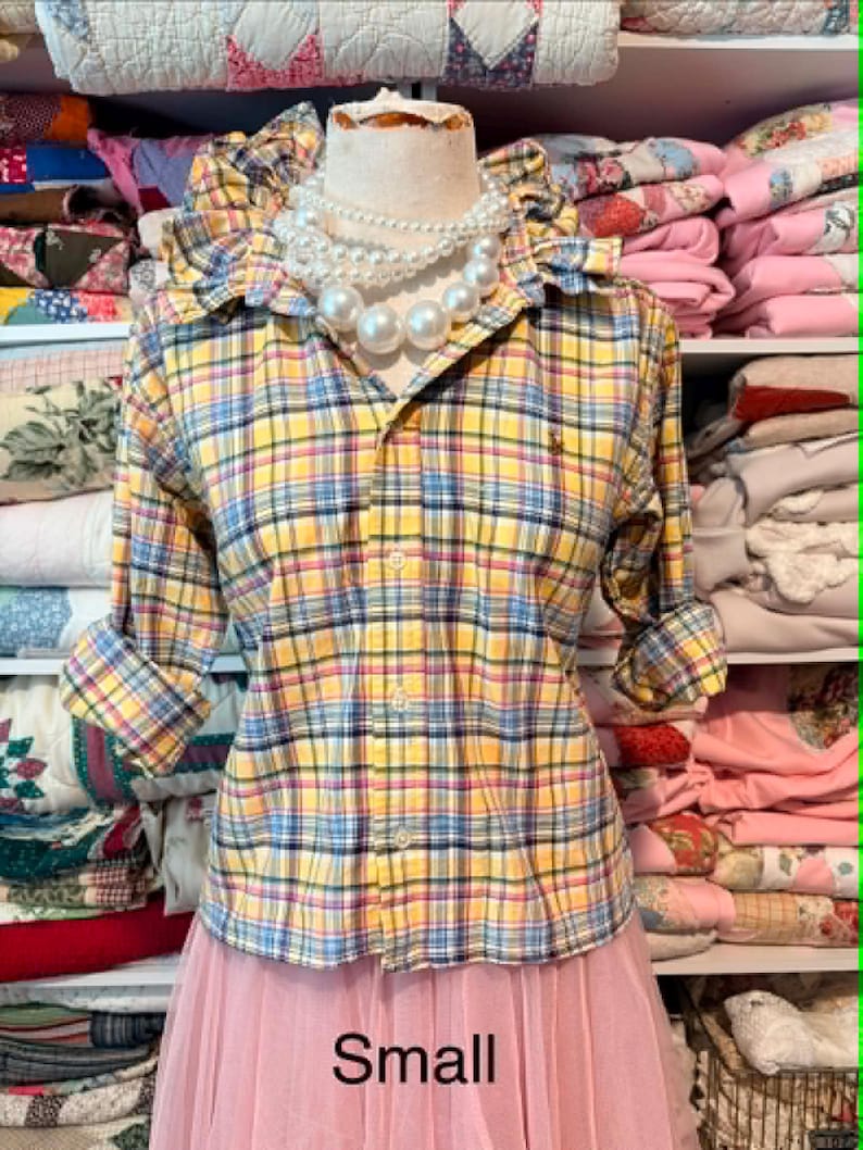 K&ouml;nnte beinhalten: Ein gelb, blau und rosa kariertes Button-Down-Hemd mit hochgekrempelten &Auml;rmeln wird auf einer Schaufensterpuppe pr&auml;sentiert. Das Hemd hat einen R&uuml;schenkragen und ein kleines Logo. Eine mehrreihige Perlenkette wird getragen. Das Wort "Small" ist auf dem rosa Rock aufgedruckt.