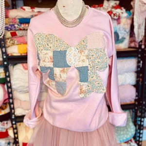 Könnte beinhalten: Ein rosa Sweatshirt mit einem großen Patchwork-Bogen auf der Vorderseite. Der Bogen besteht aus verschiedenen floralen und karierten Stoffen. Das Sweatshirt befindet sich auf einem Mannequin und wird mit einem rosa Tüllrock kombiniert.