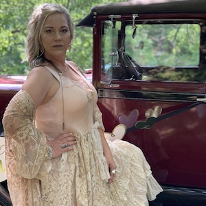 May include: A woman wearing a long, white lace dress with a sheer overlay and a lace cardigan. She is standing next to a vintage car with a red body and black trim.