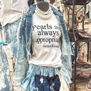 May include: A white t-shirt with the text "Pearls are always appropriate -Jackie Kennedy" printed on it. The shirt is layered over a denim skirt with a ruffled hem. The shirt is styled with a pearl necklace.