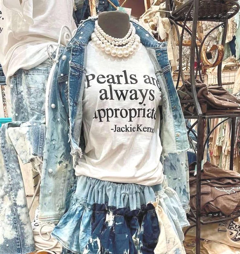 May include: A white t-shirt with the text "Pearls are always appropriate -Jackie Kennedy" printed on it. The shirt is layered over a denim skirt with a ruffled hem. The shirt is styled with a pearl necklace.