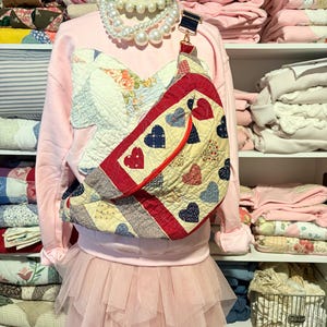 May include: A mannequin is styled in a pink sweatshirt with a patchwork design, a layered pink tulle skirt, and a pearl necklace. A quilted bag with heart and floral patterns is worn across the body. Shelves in the background are filled with folded quilts and blankets.