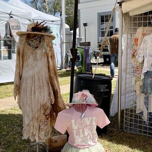 Puede incluir: Un puesto de ropa en un mercado al aire libre. Un vestido blanco con detalles de encaje est&aacute; en un maniqu&iacute;. Una camiseta rosa con el texto "Love Like Jesus" est&aacute; en un maniqu&iacute; que lleva un sombrero blanco. Un estante de ropa con una camisa blanca con estampado floral y pantalones cortos azules est&aacute; en el fondo.