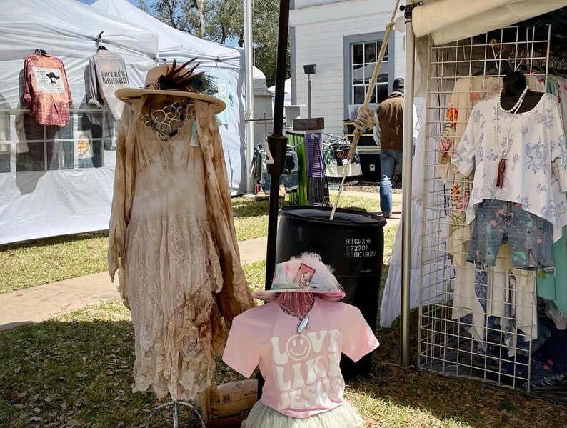 Puede incluir: Un puesto de ropa en un mercado al aire libre. Un vestido blanco con detalles de encaje est&aacute; en un maniqu&iacute;. Una camiseta rosa con el texto "Love Like Jesus" est&aacute; en un maniqu&iacute; que lleva un sombrero blanco. Un estante de ropa con una camisa blanca con estampado floral y pantalones cortos azules est&aacute; en el fondo.