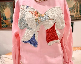 May include: A light pink long-sleeve sweatshirt featuring a large, central bow-shaped appliqué made from quilted fabric with various floral and geometric patterns in blue, red, and white. The text "Small" is printed in black in the upper left corner.