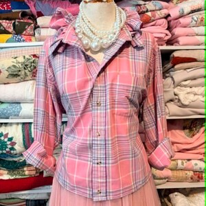 K&ouml;nnte beinhalten: Ein rosa kariertes Hemd mit R&uuml;schenkragen und hochgekrempelten &Auml;rmeln. Das Hemd wird mit einem rosa T&uuml;llrock kombiniert. Eine mehrreihige Perlenkette wird auf einer Schaufensterpuppe pr&auml;sentiert. Das Wort "Medium" ist sichtbar.
