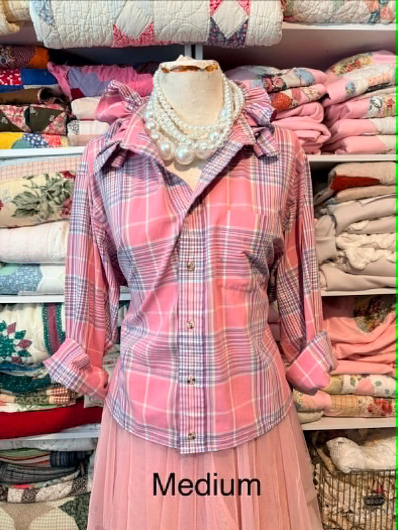 K&ouml;nnte beinhalten: Ein rosa kariertes Hemd mit R&uuml;schenkragen und hochgekrempelten &Auml;rmeln. Das Hemd wird mit einem rosa T&uuml;llrock kombiniert. Eine mehrreihige Perlenkette wird auf einer Schaufensterpuppe pr&auml;sentiert. Das Wort "Medium" ist sichtbar.