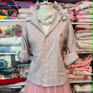 K&ouml;nnte beinhalten: Ein rot-wei&szlig; kariertes Hemd mit R&uuml;schenkragen und hochgekrempelten &Auml;rmeln, kombiniert mit einem rosa T&uuml;llrock. Eine Perlenkette schm&uuml;ckt die Schaufensterpuppe. Das Wort "Medium" ist auf den Rock gedruckt. Das Hemd wird auf einer Schaufensterpuppe pr&auml;sentiert.
