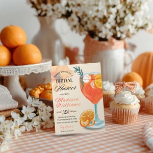 May include: A bridal shower invitation with a watercolor illustration of a cocktail with orange slices. The invitation is on a table with cupcakes, oranges, and flowers. The text reads "Bridal Shower" and the name "Madison Williams".