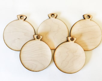 May include: Five unfinished wooden Christmas ornament cutouts. Each ornament is round with a small loop at the top for hanging. The wood has a light, natural color and visible grain.