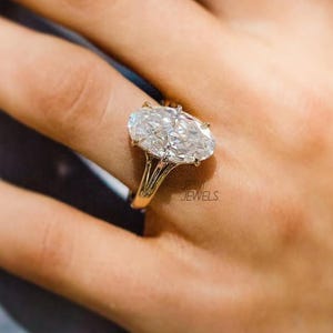 Op de afbeelding: Een gouden ring met een grote, ovaal geslepen diamant. De heldere, sprankelende diamant is gezet op een delicate gouden band. De ring zit om een vinger, met de merknaam "JESVI JEWELS" zichtbaar.