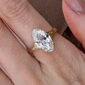 Op de afbeelding: Een gouden ring met een ovaal geslepen diamant. De diamant is het middelpunt en reflecteert het licht. De ringband heeft een ingewikkeld ontwerp. De ring zit om een vinger, met het Jesvi Jewels-logo zichtbaar.