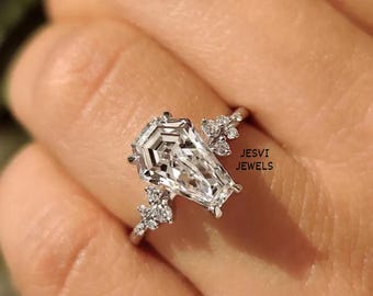 May include: A silver-toned ring featuring a large, coffin-shaped centre stone and smaller accent stones clustered on the band. The text JESVI JEWELS is visible on the right side of the ring. The design includes a polished metal band.