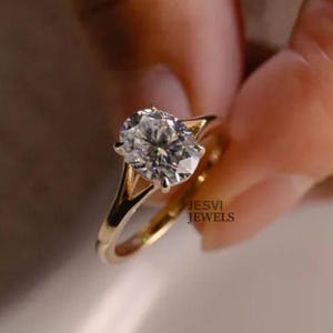 Könnte beinhalten: Ein goldener Ring mit einem großen, oval geschliffenen Diamanten. Der klare Diamant funkelt in einer Vier-Krappen-Fassung. Der Ring ist aus poliertem Gold. Der Text "JESVI JEWELS" ist auf dem Ring zu sehen.
