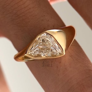 Könnte beinhalten: Ein goldener Ring mit einem großen, dreieckigen, klaren Edelstein. Der Ring hat ein glattes, poliertes Band und eine Lünettenfassung. Ein auffälliges Schmuckstück.