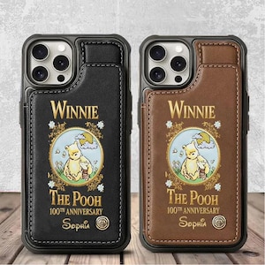 May include: Two phone cases featuring a Winnie the Pooh design. One case is black, the other is brown. Both cases have a card holder and a circular illustration of Winnie the Pooh and Piglet. The text "Winnie The Pooh 100th Anniversary" is printed on the cases.