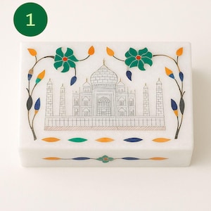 May include: White marble box with a Taj Mahal design. The box features a detailed black and white illustration of the Taj Mahal, with colorful floral and leaf accents in green, orange, and blue. The box is rectangular and has a lid.