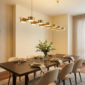 May include: A modern dining room with a dark wood table set for a meal. Above the table hangs a gold-toned linear chandelier. A round mirror and a vase of flowers add to the room's decor. The room has a neutral color palette.