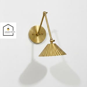 May include: A brass wall sconce with an adjustable arm and a pleated shade. The sconce is mounted on a round backplate and casts a shadow on the white wall. The logo "GOLDEN SPOON" is in the lower left corner.