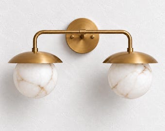 May include: A wall-mounted light fitting featuring a horizontal brass arm attached to a circular backplate. Two downward-facing brass domes hold spherical white marble globes with subtle brown veining, creating a balanced lighting design.