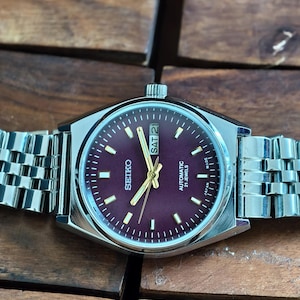 May include: A Seiko automatic wristwatch with a burgundy dial and silver-tone hands and markers. The watch features a stainless steel case and a silver link bracelet. The day and date are displayed at the 3 o'clock position.