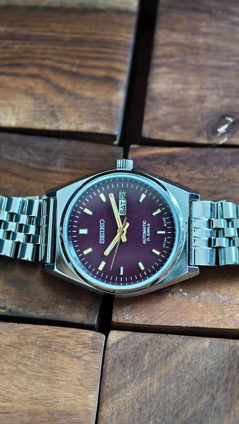 May include: A Seiko automatic wristwatch with a burgundy dial and silver-tone hands and markers. The watch features a stainless steel case and a silver link bracelet. The day and date are displayed at the 3 o'clock position.