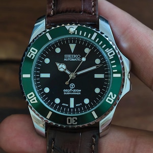 K&ouml;nnte beinhalten: Eine Seiko Automatic Submariner Uhr mit schwarzem Zifferblatt und gr&uuml;ner L&uuml;nette. Die Uhr hat ein braunes Lederarmband. Das Zifferblatt zeigt den Text "660ft=200m Submariner".