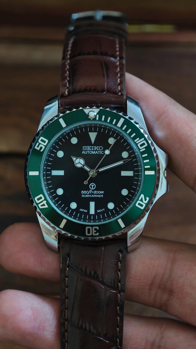 K&ouml;nnte beinhalten: Eine Seiko Automatic Submariner Uhr mit schwarzem Zifferblatt und gr&uuml;ner L&uuml;nette. Die Uhr hat ein braunes Lederarmband. Das Zifferblatt zeigt den Text "660ft=200m Submariner".