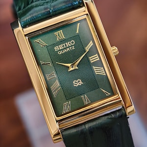 May include: A rectangular Seiko Quartz watch with a gold-tone case and a green dial. The dial features gold Roman numerals and hands. The watch has a green leather strap.
