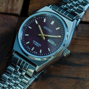 May include: A stainless steel Seiko automatic wristwatch with a burgundy dial. The watch features gold hands and markers, a day-date display, and a silver link bracelet. The watch is displayed on a wooden surface.