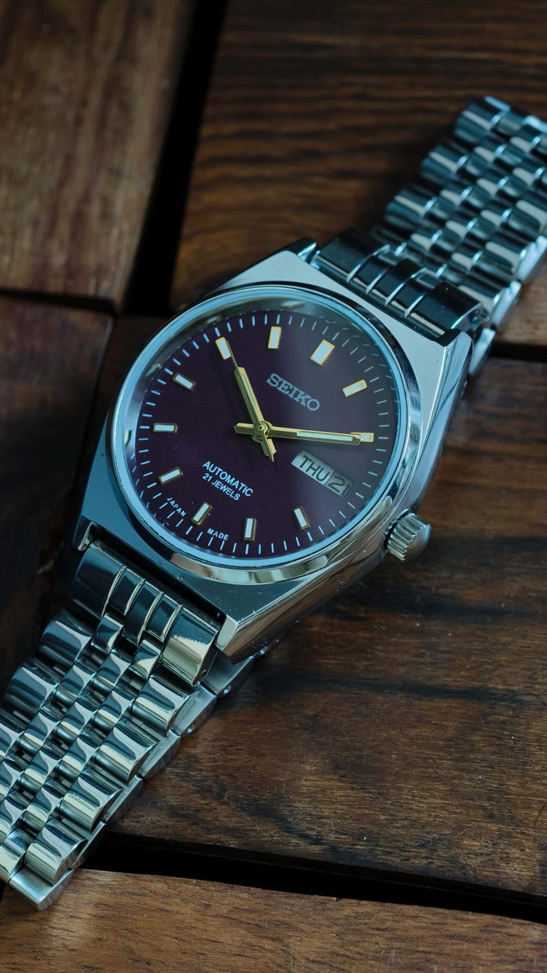 May include: A stainless steel Seiko automatic wristwatch with a burgundy dial. The watch features gold hands and markers, a day-date display, and a silver link bracelet. The watch is displayed on a wooden surface.