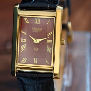 Peut inclure: Une montre Seiko Quartz dorée avec un cadran bordeaux et des chiffres romains dorés. La montre a des aiguilles dorées et un bracelet en cuir noir. Le cadran de la montre porte le logo Seiko Quartz.
