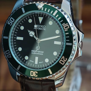 K&ouml;nnte beinhalten: Nahaufnahme einer Seiko Automatik-Uhr mit schwarzem Zifferblatt und gr&uuml;ner L&uuml;nette. Die Uhr hat ein braunes Lederarmband. Auf dem Zifferblatt steht "SEIKO AUTOMATIC" und "SUBMARINER".
