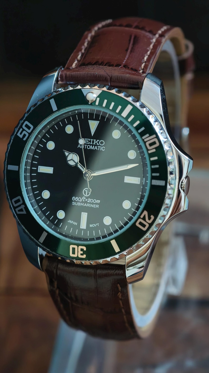 K&ouml;nnte beinhalten: Nahaufnahme einer Seiko Automatik-Uhr mit schwarzem Zifferblatt und gr&uuml;ner L&uuml;nette. Die Uhr hat ein braunes Lederarmband. Auf dem Zifferblatt steht "SEIKO AUTOMATIC" und "SUBMARINER".
