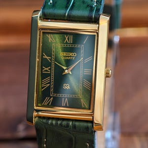 May include: A rectangular gold-toned Seiko Quartz watch with a green dial and Roman numeral hour markers. The watch has a green leather band. The watch face displays the Seiko logo and the word "Quartz".