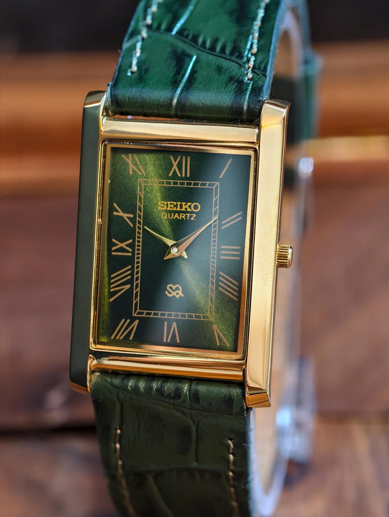 May include: A rectangular gold-toned Seiko Quartz watch with a green dial and Roman numeral hour markers. The watch has a green leather band. The watch face displays the Seiko logo and the word "Quartz".