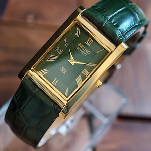 May include: A rectangular Seiko Quartz watch with a green face and gold hands and markers. The watch has a gold case and a green leather strap. The watch face displays Roman numerals. A classic timepiece.