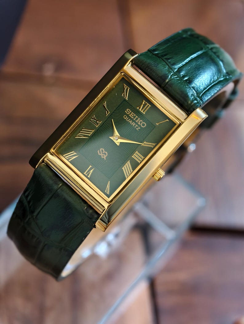 May include: A rectangular Seiko Quartz watch with a green face and gold hands and markers. The watch has a gold case and a green leather strap. The watch face displays Roman numerals. A classic timepiece.