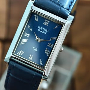 May include: A rectangular Seiko Quartz watch with a navy blue face and silver case. The watch has Roman numeral hour markers and a navy blue leather strap. The watch face displays the Seiko Quartz logo.