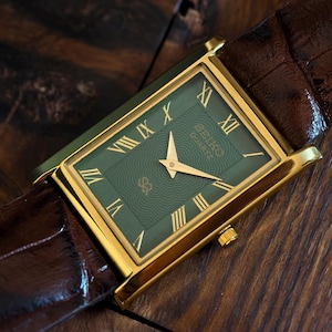 May include: A rectangular gold-toned wristwatch with a green face and Roman numeral hour markers. The watch has a brown leather strap. The watch face features a wave pattern and a logo. The watch is set against a wooden surface.