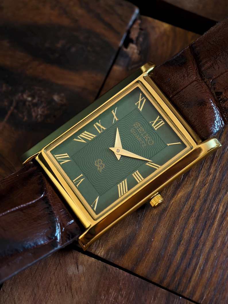May include: A rectangular gold-toned wristwatch with a green face and Roman numeral hour markers. The watch has a brown leather strap. The watch face features a wave pattern and a logo. The watch is set against a wooden surface.
