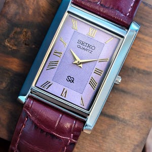 Könnte beinhalten: Eine rechteckige Seiko Quartz-Uhr mit lavendelfarbenem Zifferblatt und goldenen römischen Ziffern. Die Uhr hat ein silbernes Gehäuse und ein bordeauxfarbenes Lederarmband. Die Uhr wird auf einer Holzoberfläche präsentiert.